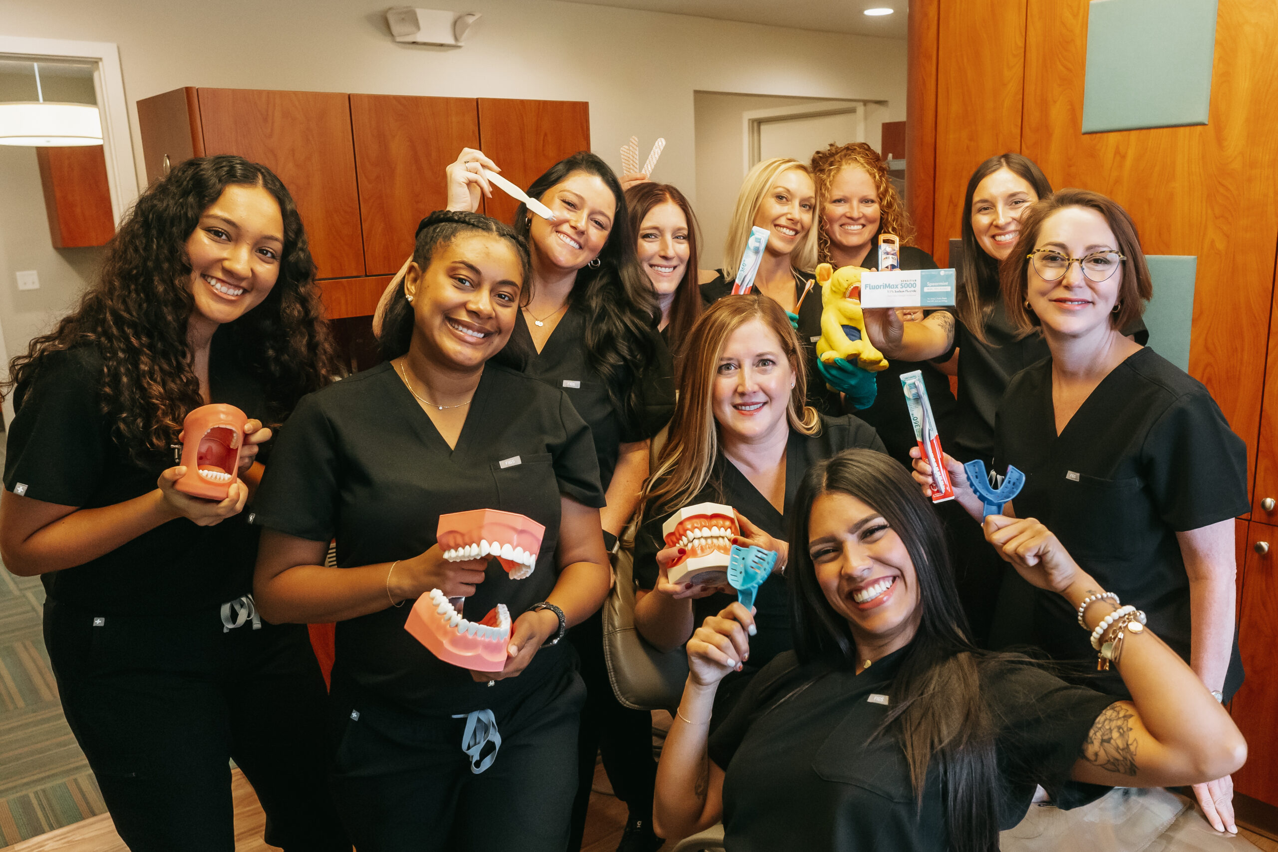 A cheerful team of dental hygienists and assistants in black uniforms smile while holding dental tools, toothbrushes, and model teeth