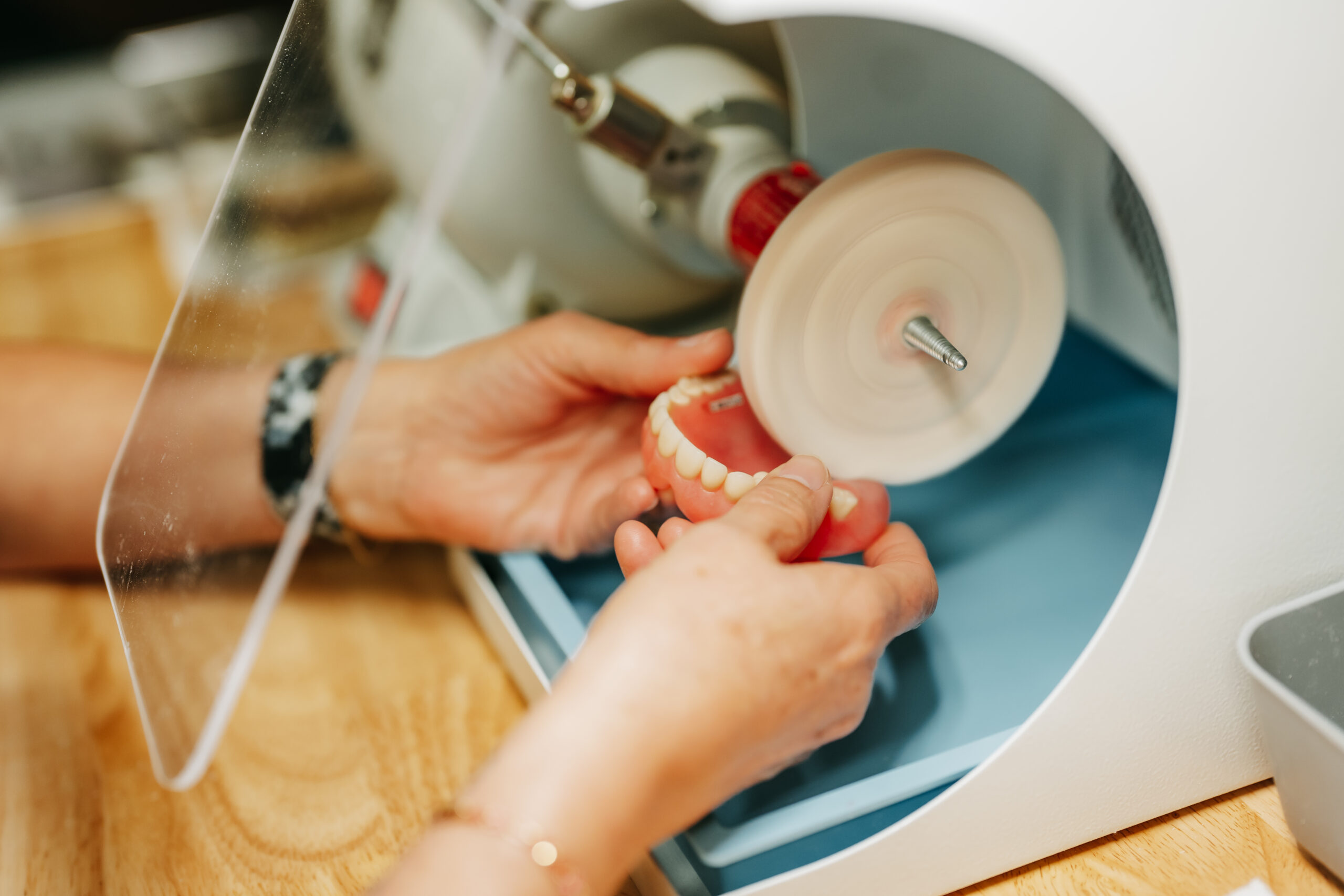 A close-up view of an expert dental technician carefully polishing a set of dentures using a rotating buffing wheel in a dental lab. The technician's hands hold the dentures steadily while the polishing machine smooths the surface.