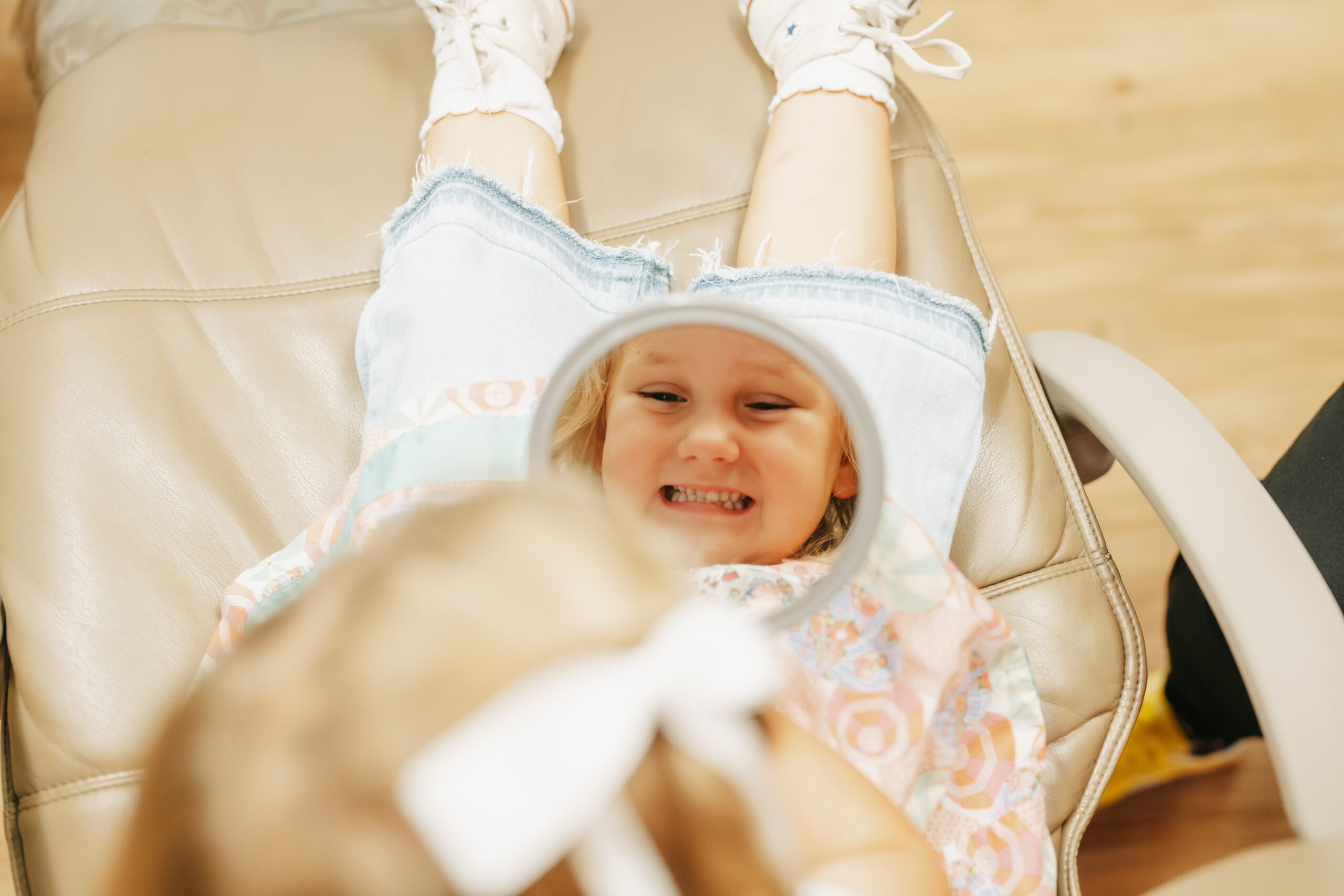 Happy young child lying back in a dental chair, joyfully looking at her reflection in a mirror and showing off her bright, healthy smile