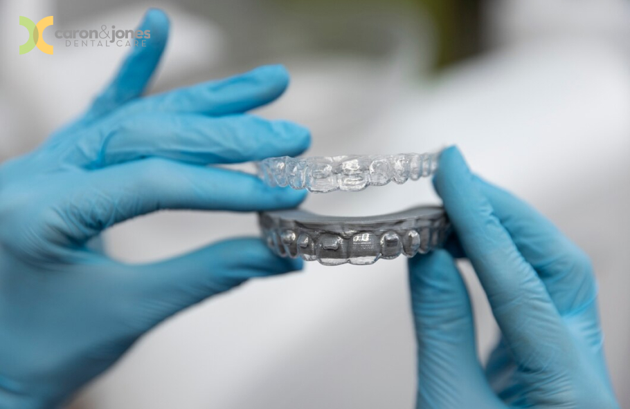 A close-up image of a person holding clear aligners between their fingers. The focus is on the transparent aligners, showcasing their smooth, custom-fit design and flexibility, with a blurred background to emphasize the aligners.